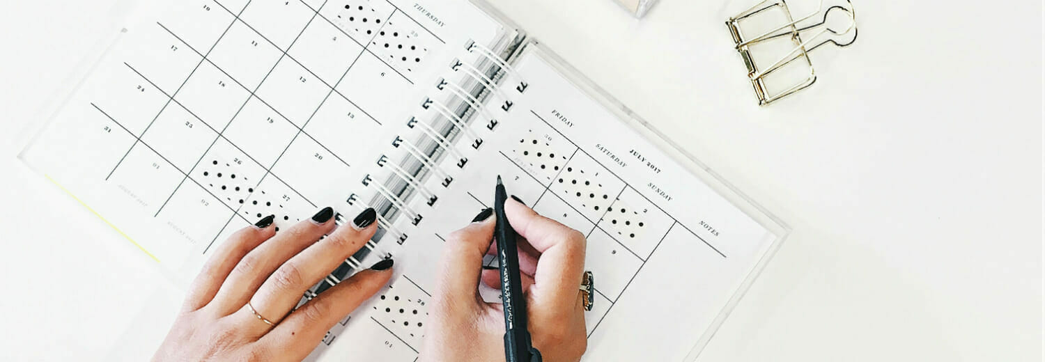 Overhead shot of woman's hands writing in weekly planner on white tabletop