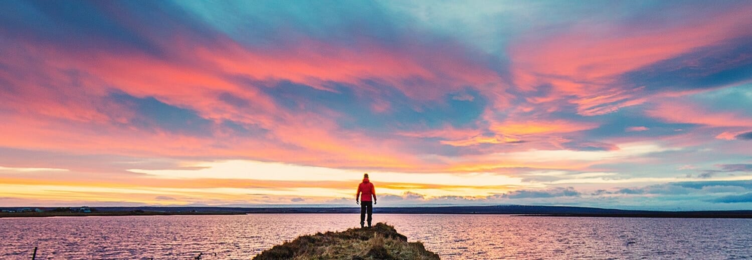 Man in a red jacket standing on a rock gazing into a pink sunset