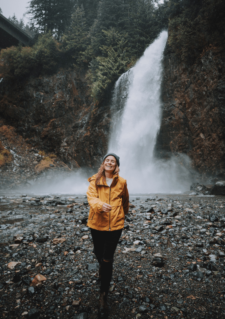 Person in front of a foreest wth a waterfall smiling and walking towards the camera